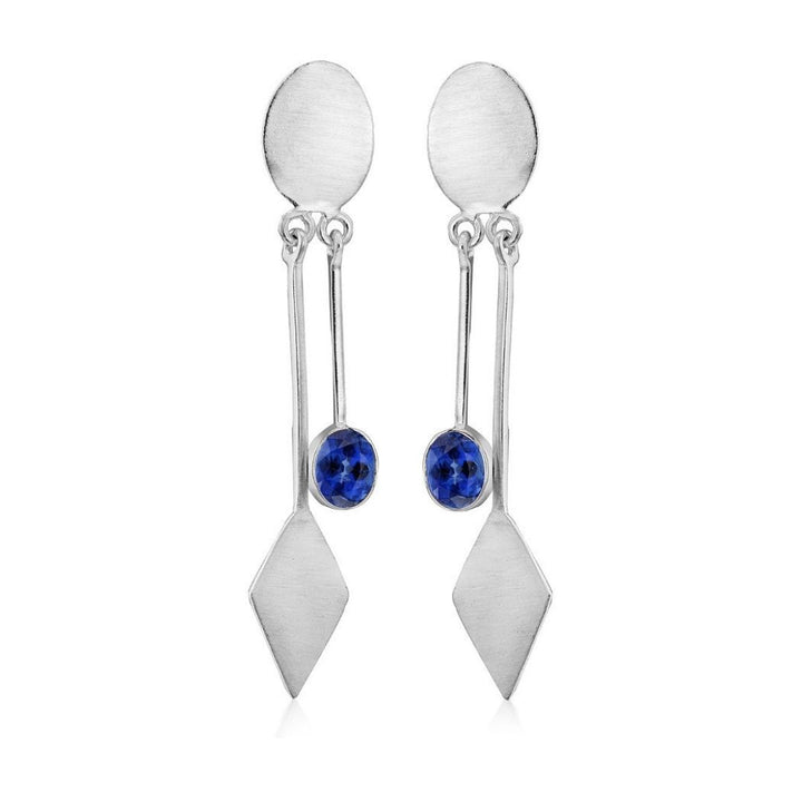 Discover modern elegance with Gallardo & Blaine Designs' Karo Earrings, silver dangle drops featuring oval studs and a sleek bar leading to a diamond-shaped drop. A small blue gemstone near the bottom enhances these chic accessories.