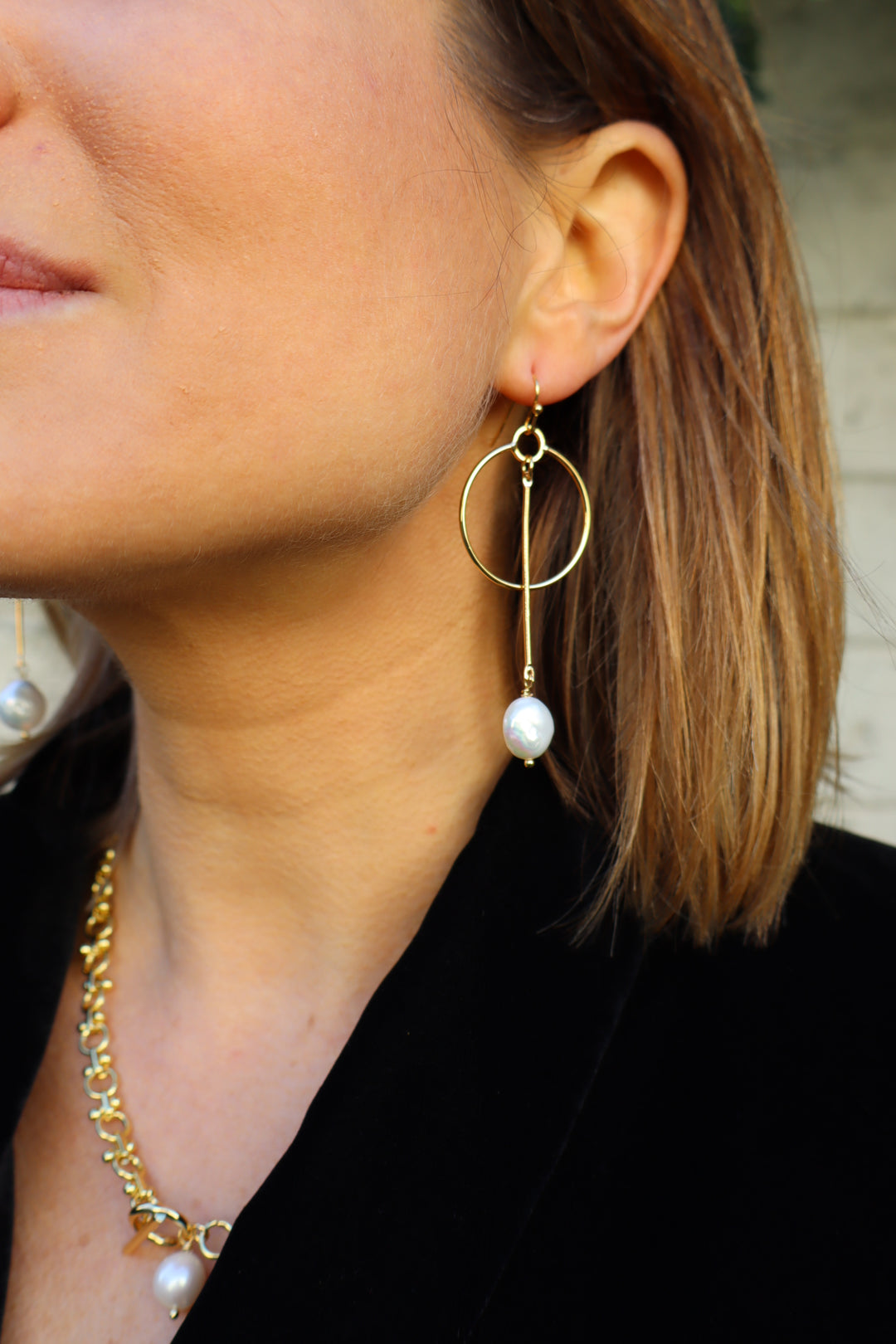 Close-up of a person wearing Kybalion Jewellery’s Kea Earrings with freshwater pearl drops, paired with a gold chain necklace. The handmade Dublin jewelry pops against their short brown hair and black top.