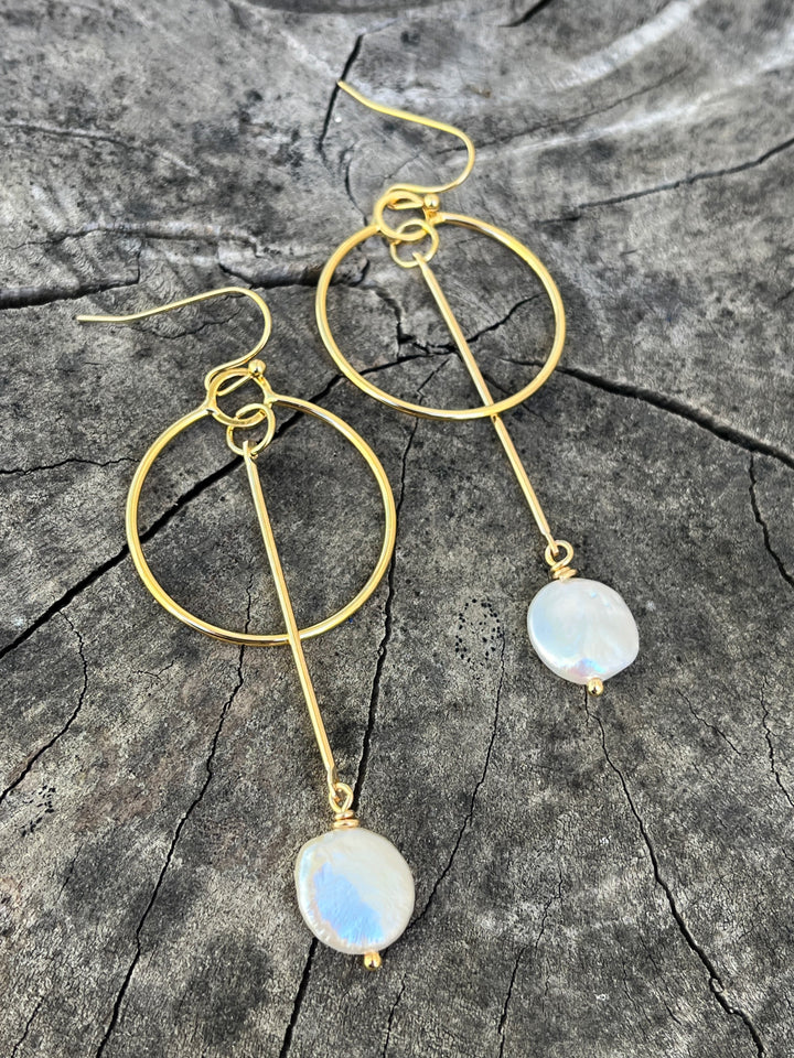 The Kea Earrings with Freshwater Pearl Drops by Kybalion Jewellery feature gold-plated hoops with vertical bars and iridescent pearls, beautifully displayed on textured gray wood—elegant handmade Dublin jewelry.