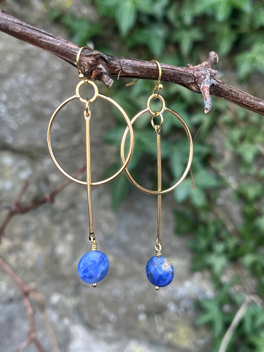 Kea Earrings with Sodalite Drop by Kybalion Jewellery: gold plated earrings with a vertical bar and sodalite disc, shown on a tree branch against a blurred green leafy background.