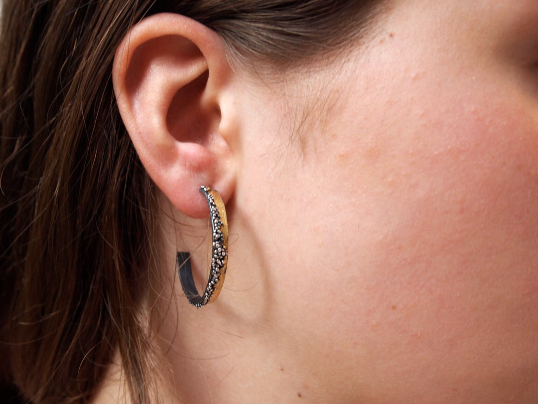 Close-up of a person's ear with straight brown hair, wearing a textured silver Etrusc Hoop Earring by MarCucurella. Smooth skin highlights the earring, available in various sizes from the brand's Etrusc collection.