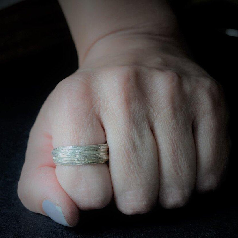 A close-up of a hand with a sterling silver ring on the middle finger. The hand is resting on a dark surface, and the fingers are slightly curled inward. The lighting focuses on the Mitsuro Silver Ring by Abbie Dixon, casting a shadow on the surface.