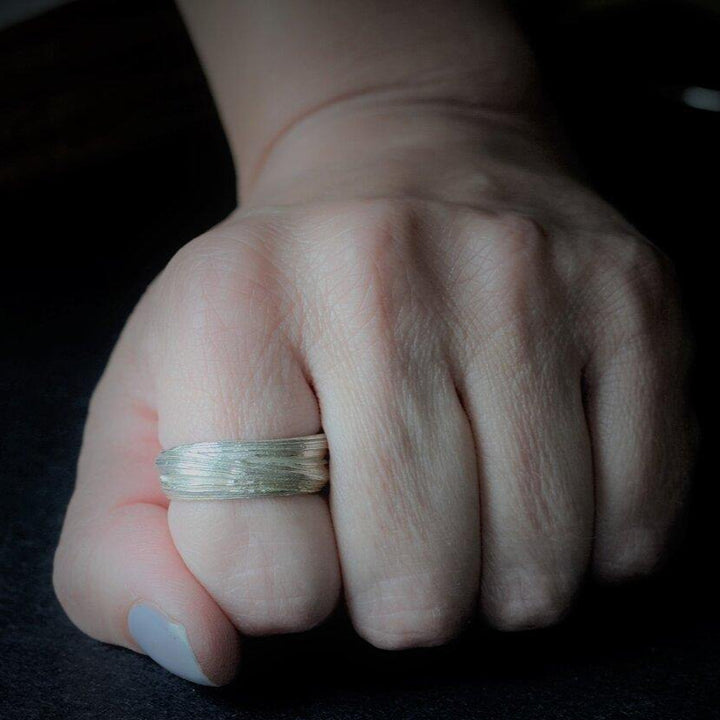 A close-up of a hand with a sterling silver ring on the middle finger. The hand is resting on a dark surface, and the fingers are slightly curled inward. The lighting focuses on the Mitsuro Silver Ring by Abbie Dixon, casting a shadow on the surface.