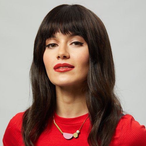 A woman with long dark hair and bangs, in a bright red top and bold lipstick, wears the Shock of Grey Multishape Plus Necklace in Dirty Pink & Sage; she smiles softly against a plain light background.