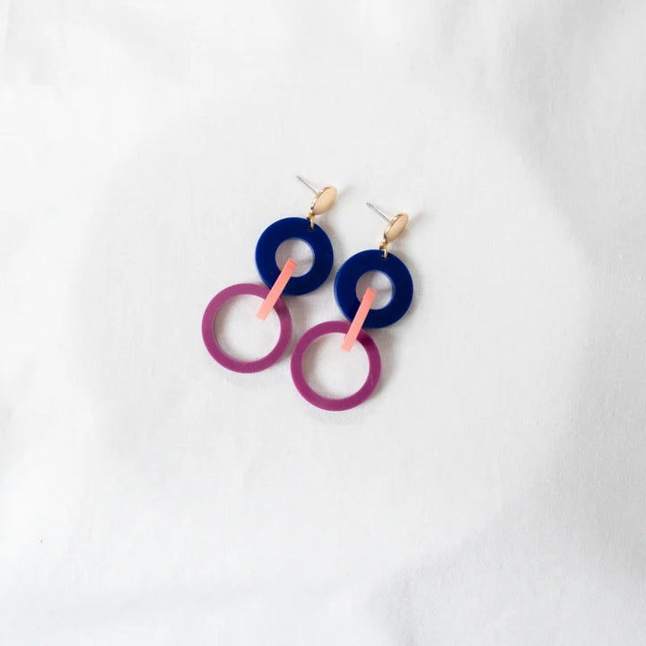 A pair of Studio Nok Nok Navy, Pink & Magenta Acrylic Fire Earrings laid on a white surface. Each earring consists of two interlinked round loops, one dark blue and one light purple, connected by a pink vertical bar made from recycled acrylic. The earrings have gold-tone studs at the top.