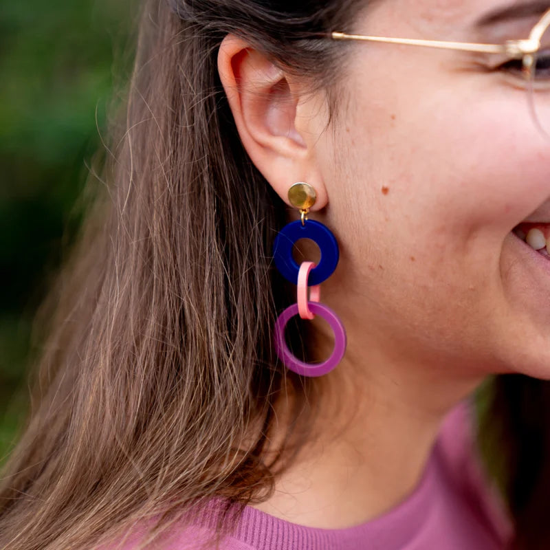 A close-up side view of a smiling person wearing Studio Nok Nok's Navy, Pink & Magenta Acrylic Fire Earrings. The person has shoulder-length brown hair, glasses, and is dressed in a pink top. The background is blurred greenery.