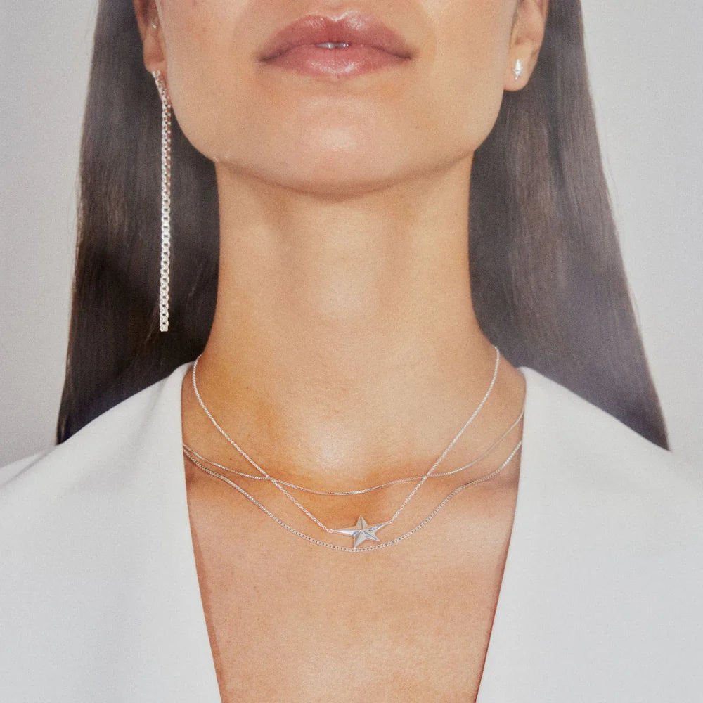 A woman wearing a white top and a long earring showcases three Jane Koenig 925 sterling silver necklaces, including the Night Star Necklace, which features a star pendant, against a neutral background.