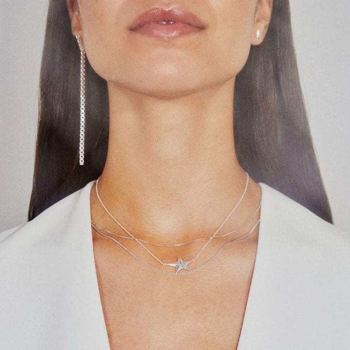 A woman wearing a white top and a long earring showcases three Jane Koenig 925 sterling silver necklaces, including the Night Star Necklace, which features a star pendant, against a neutral background.