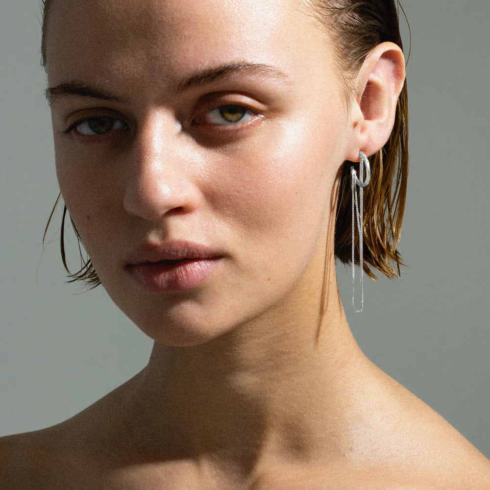 A woman with wet, slicked-back hair wears the Jane Koenig Orbis Chain Hoop—sterling silver double hoops with a delicate chain. With minimal makeup, she gazes calmly at the camera against a neutral background, expressing modern jewelry style.