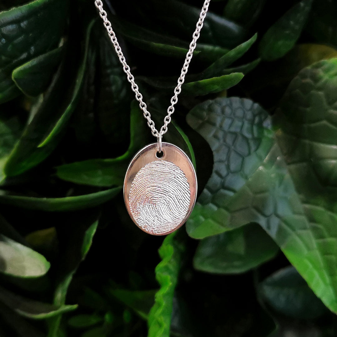 Displayed against lush green leaves, the Engraved Fingerprint Pendant by Personalised Charms is a silver necklace featuring a customized round pendant with a unique fingerprint design.