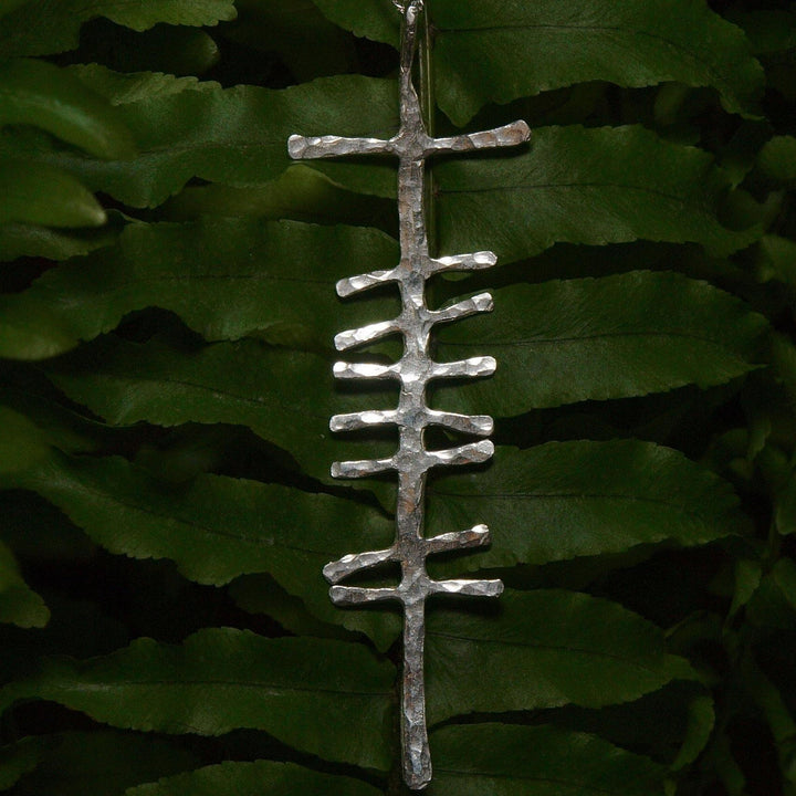 A thin, silver pendant featuring a unique, abstract design hangs against the backdrop of lush, green fern leaves. The Love (Grá) Large Rustic Pendant Silver’s vertical structure resembles an artistic ladder or runic symbol, reminiscent of ancient Ogham writing, from Ogham Treasure.