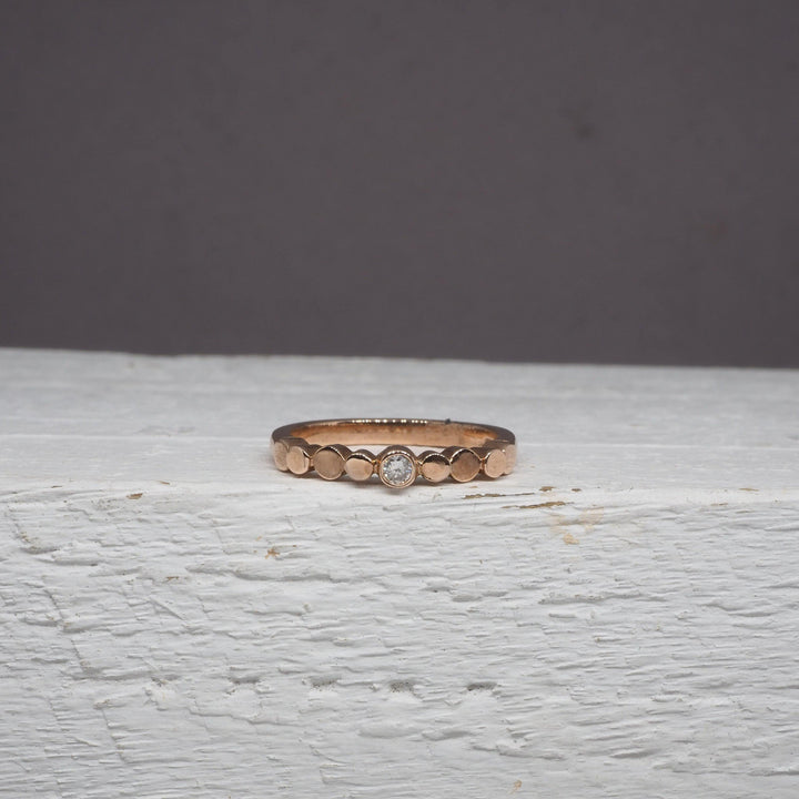 A delicate Flow Pebble Ring 9ct Rose Gold with Diamond by Miriam Wade rests on a white, wooden surface against a plain gray background. This exquisite piece is made to order.