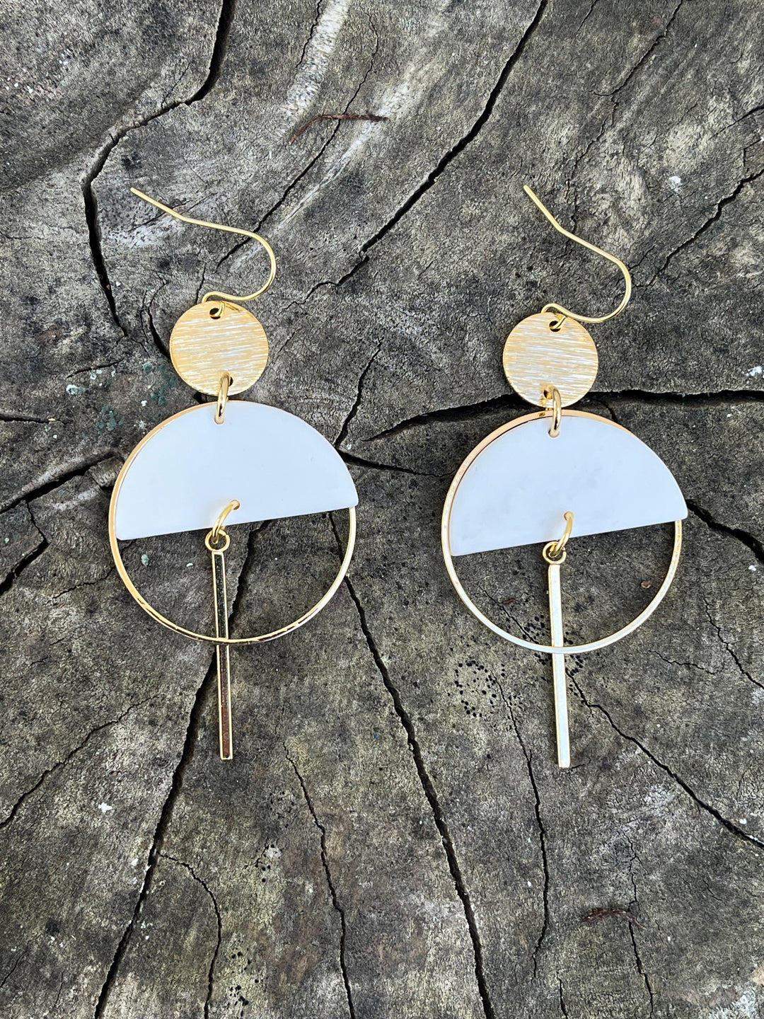 The Sinead Earrings by Kybalion Jewellery feature modern gold-plated circular and semicircular shapes with hanging bars and colorful polymer half moons, shown on a rustic wooden surface.