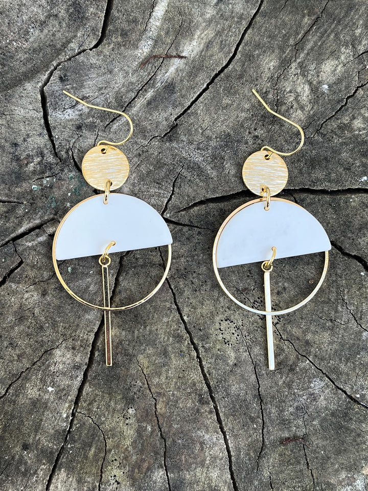 The Sinead Earrings by Kybalion Jewellery feature modern gold-plated circular and semicircular shapes with hanging bars and colorful polymer half moons, shown on a rustic wooden surface.