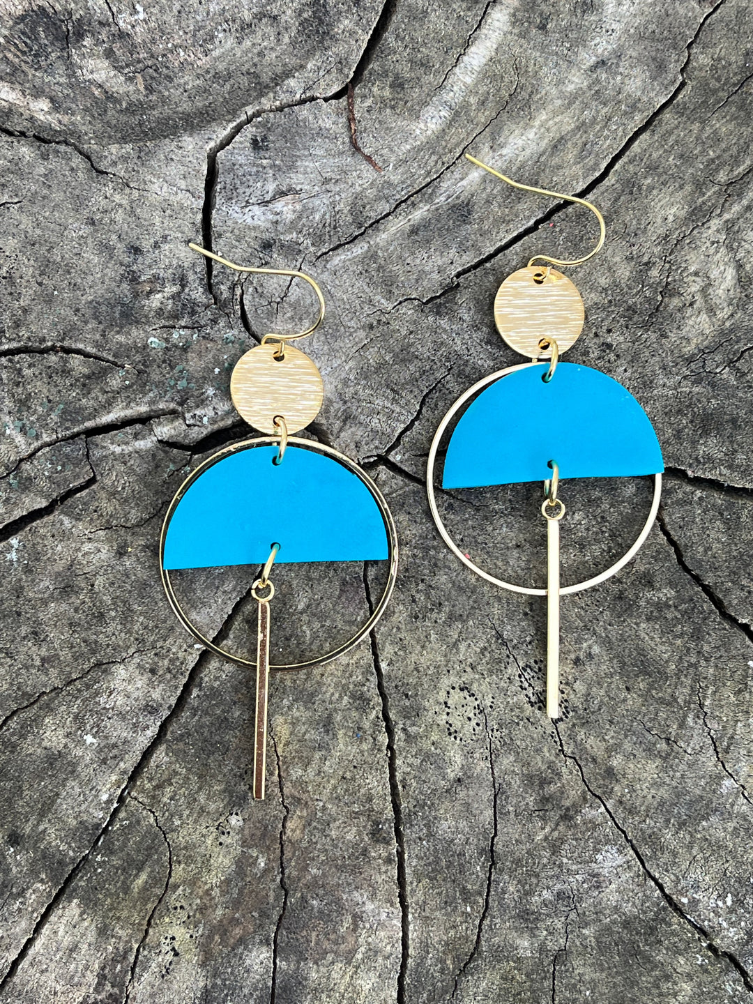 The Sinead Earrings by Kybalion Jewellery feature gold-plated hoops, textured discs, bright blue polymer half moons, and vertical gold bars, beautifully displayed on a cracked wooden surface.