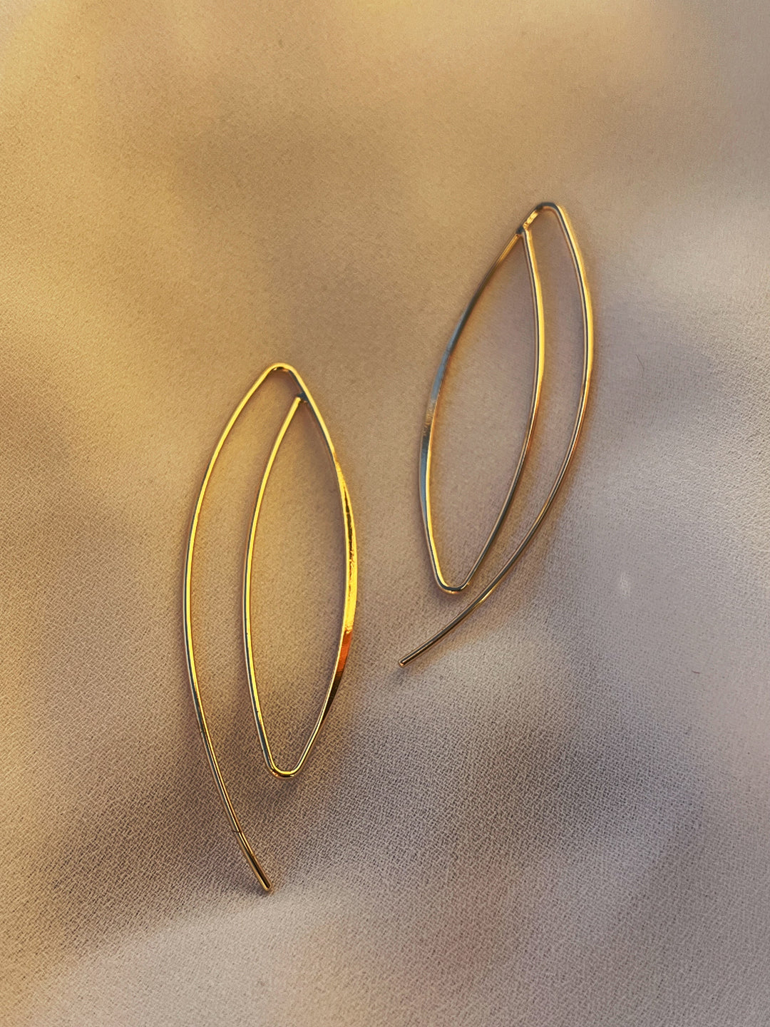 JoJo Earrings by Kybalion Jewellery, crafted in a gold leaf shape and gold plated, rest elegantly on a soft, light-toned surface, as gentle lighting casts a warm, subtle glow around them.