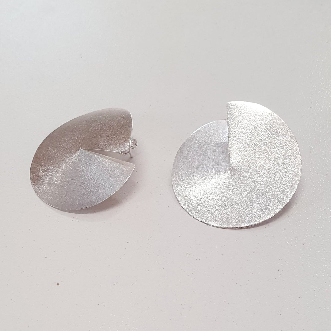 Two matte silver Split Disc Earrings by Georgia Charal, handmade in Athens and featuring abstract, folded disc shapes with a textured surface, are displayed on a light background.
