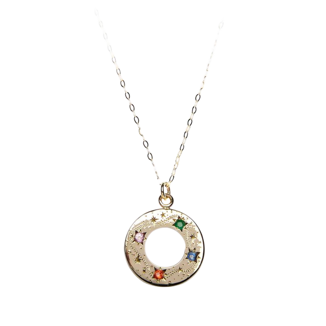 A delicate 9ct yellow gold necklace, the Shooting Star Rainbow Pendant 9ct Yellow Gold from Cosmic Boulevard, features a circular pendant adorned with small, star-shaped engravings reminiscent of shooting stars and four colored gemstones: green (emerald), pink, orange, and blue (sapphires). The chain is thin and elegant, with a simple link design.