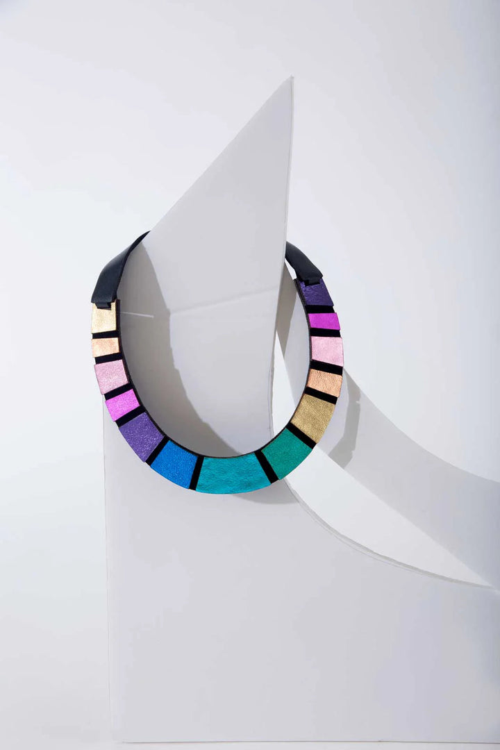The Rainbow Leather Necklace by Iskin Sisters is showcased against a minimalist, white abstract background. This vibrant piece features multicolored geometric shapes in shades of pink, gold, blue, green, and black on lightweight Argentinian leather, creating a striking visual effect.