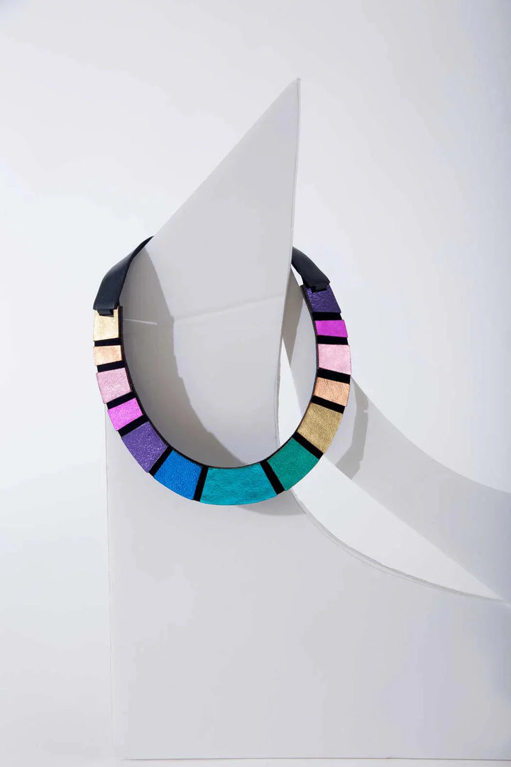 The Iskin Sisters Rainbow Round Leather Necklace features vibrant blue, purple, pink, green, and gold segments, showcased on a white abstract background with angular shapes and shadows.