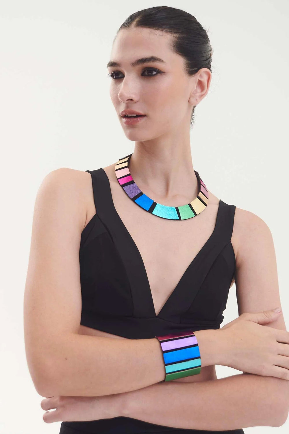 A person with dark hair is wearing a sleeveless black outfit, accessorized with the vibrant Iskin Sisters Rainbow Leather Necklace and matching bracelet, featuring rectangular segments in hues of pink, blue, green, purple, and gold. They pose against a plain white background.