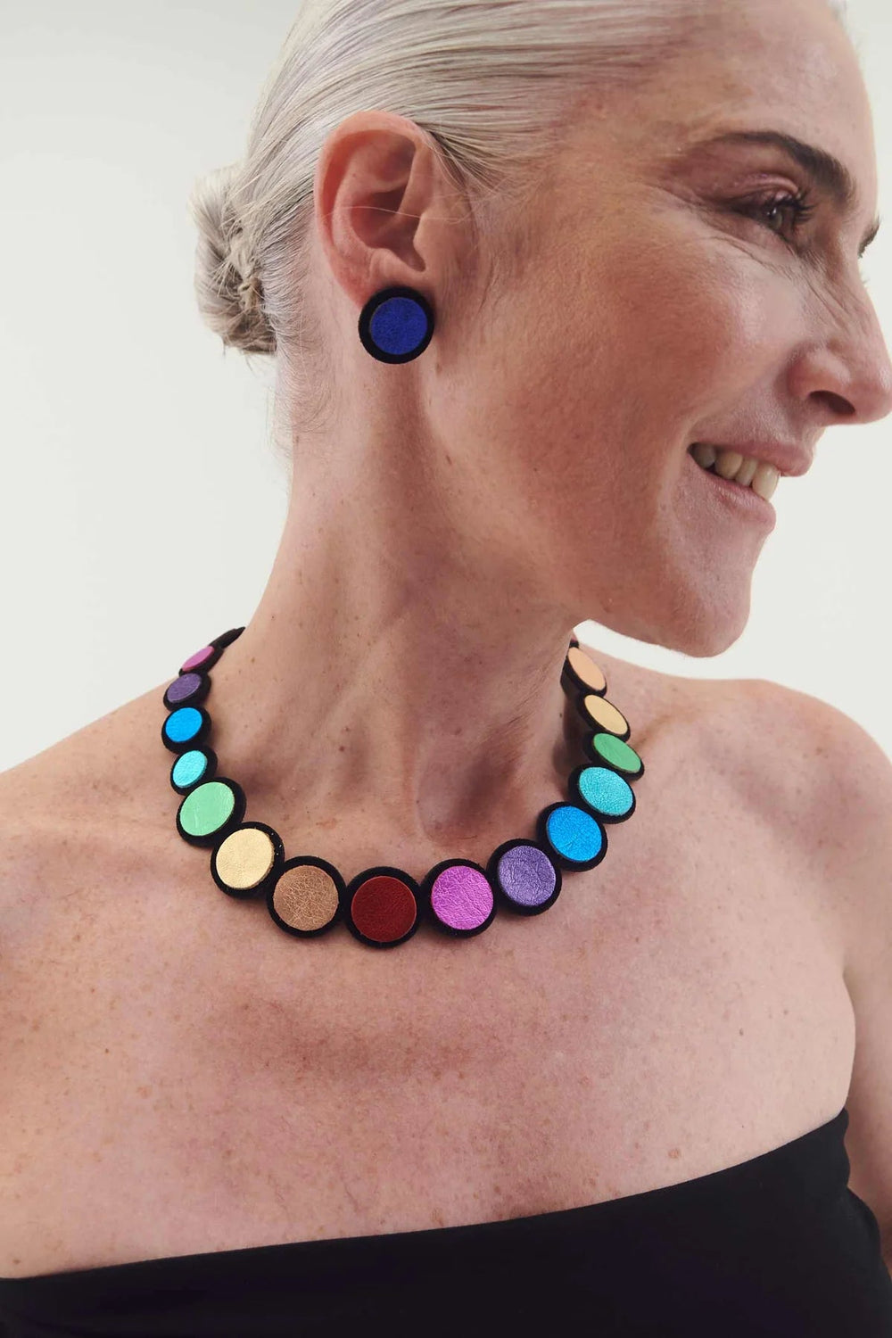 A woman with short, pulled-back gray hair is smiling in a strapless black top. She is wearing the vibrant Rainbow Round Leather Necklace and matching earrings from Iskin Sisters, featuring beads in shades of blue, purple, red, and gold. The background is plain white.