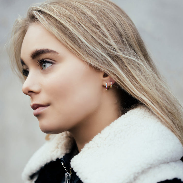 A young woman with long blonde hair and light eyes looks left, wearing a black jacket with a white shearling collar and Sif Jacobs Ellera Medio Hoop Earrings. The background is softly blurred.