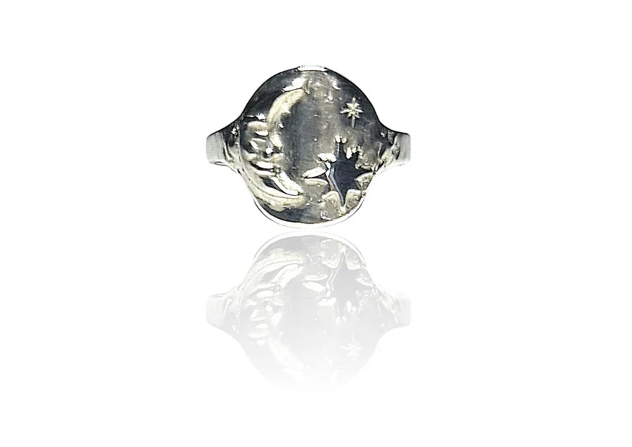 The Pale Raven "A Sailor's Dream" Silver Signet Ring features an engraved crescent moon, star, and cloud on a polished face with a relic-inspired look, reflected elegantly on a white surface.