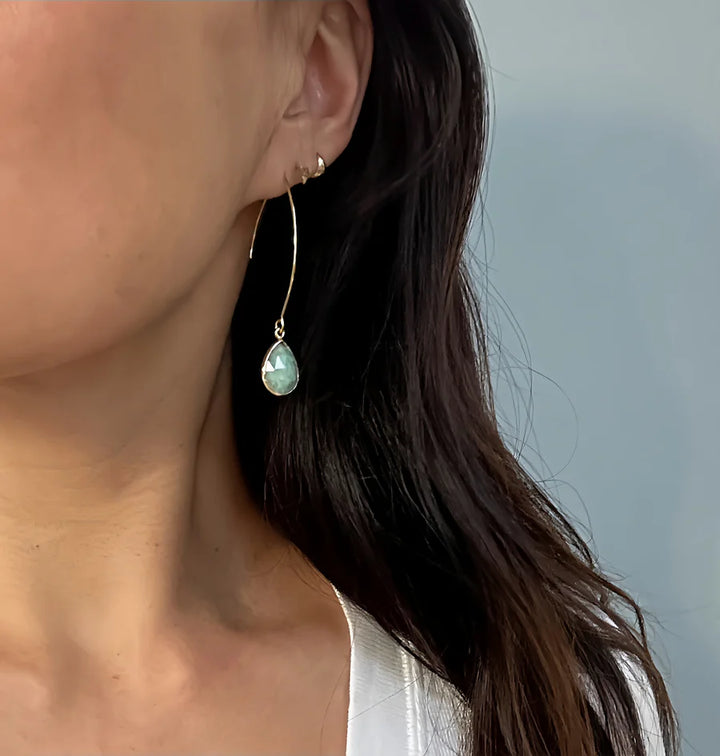A close-up shows a woman with long brown hair in a white top wearing Aria V's Sea Foam Earrings—long, handcrafted gold hoops with teardrop-shaped light blue amazonite stones—her ear and the softly blurred background in focus.