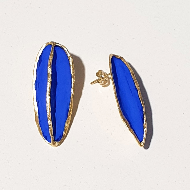 Georgia Charal’s Leaf Earrings in cobalt blue feature bold enamel, irregular gold-plated edges, and unique asymmetrical shapes—one oval and one elongated—shown on a light background.