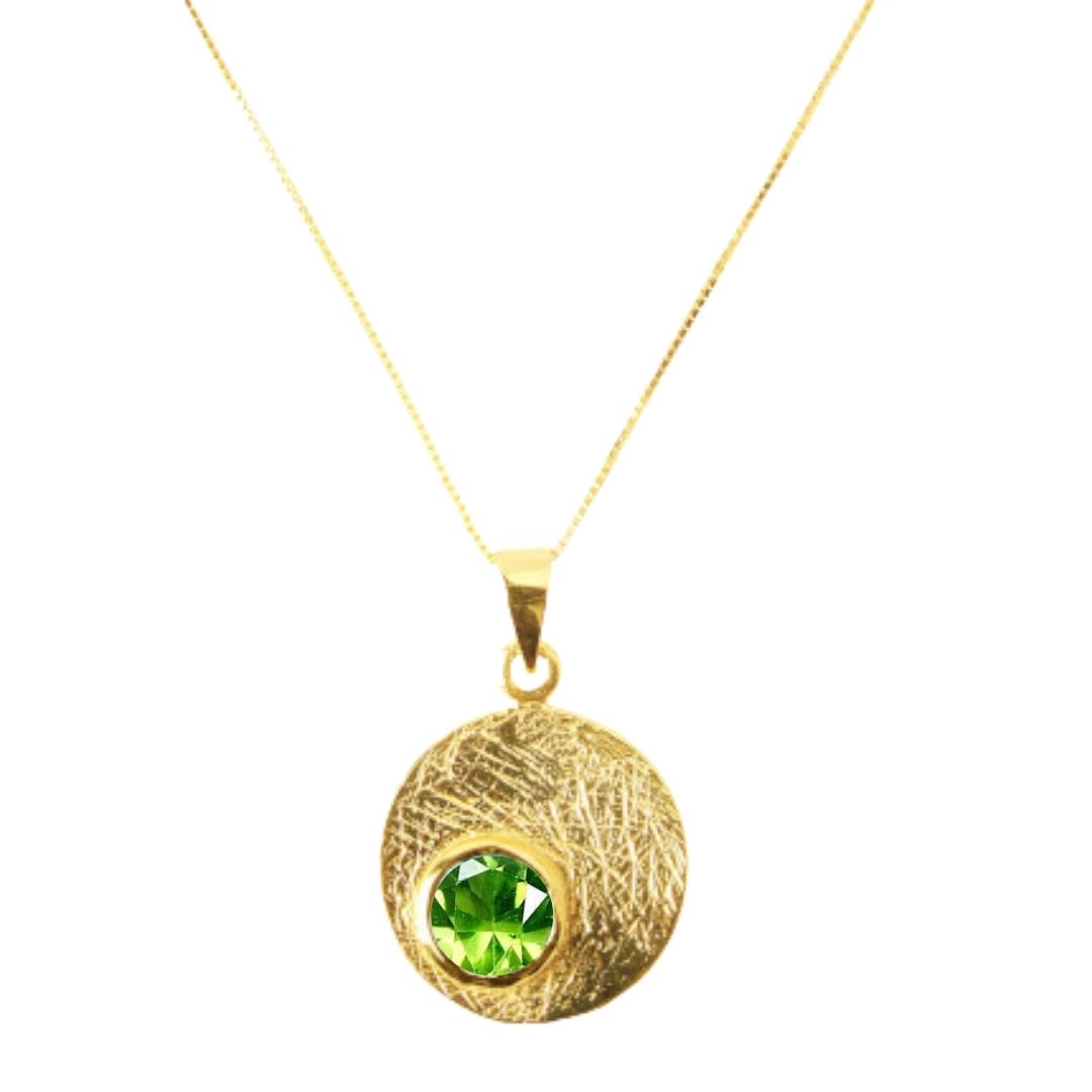 The Selene Necklace Gold - Small by Gallardo & Blaine Designs features a circular, textured gold-plated pendant with a bright green gemstone at its center, suspended from a fine gold chain.