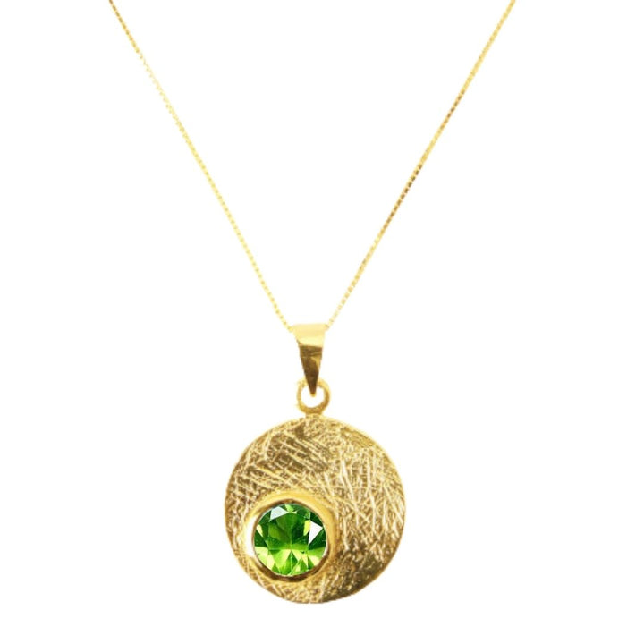 The Selene Necklace Gold - Small by Gallardo & Blaine Designs features a circular, textured gold-plated pendant with a bright green gemstone at its center, suspended from a fine gold chain.