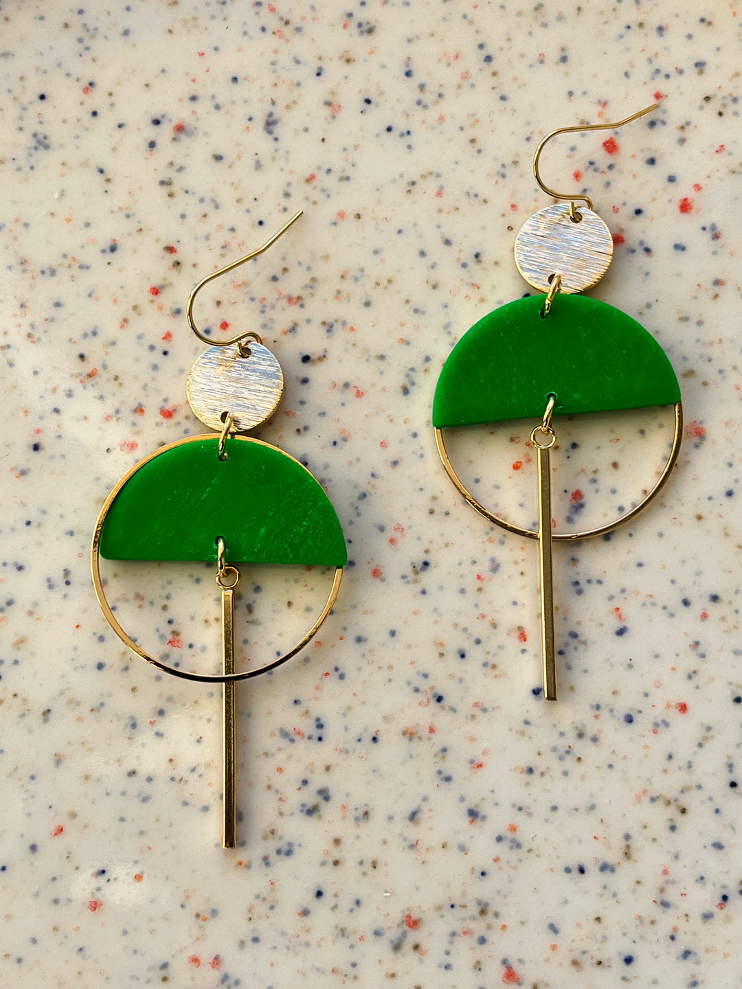The Sinead Earrings by Kybalion Jewellery showcase silver round studs, vibrant polymer half moons, gold hoops, and vertical bars in modern gold-plated style—available in various colours, displayed on a speckled beige background.