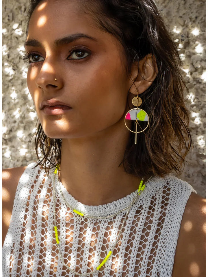 A woman with short, wavy hair and dewy skin wears a white crochet top, layered necklaces, a nose stud, and Kybalion Jewellery’s Sinead Earrings in neon yellow and pink. Dappled sunlight filters across her face and background.