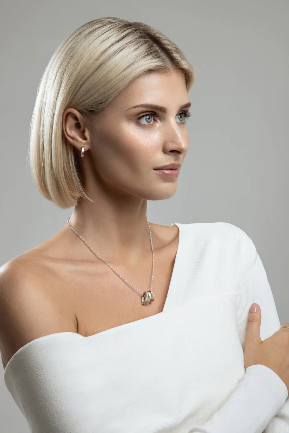 A woman with short blonde hair is wearing a white off-the-shoulder top and Talún Studs by Lynsey de Burca. She has a Sterling silver pendant necklace and her arms are crossed in front of her as she gazes to the side. The background is grey, creating a soft, minimalist look that highlights her handmade jewelry.