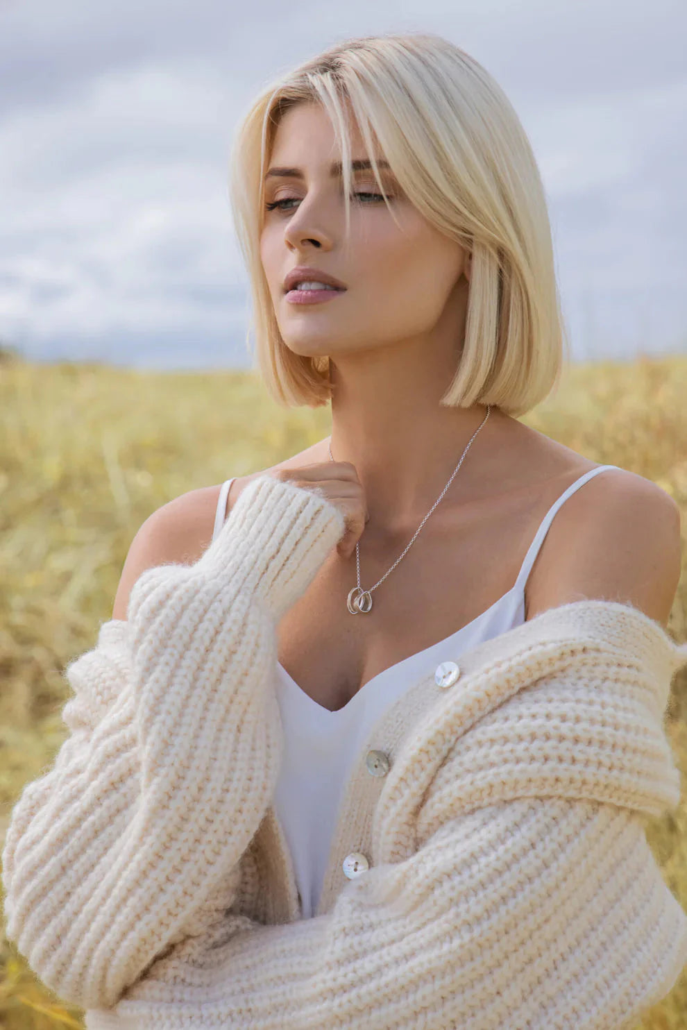 A woman with short blond hair in a white dress and chunky knit cardigan stands in a Connemara field, gazing thoughtfully as sunlight illuminates her Lynsey de Burca Talún Triple Neckpiece.