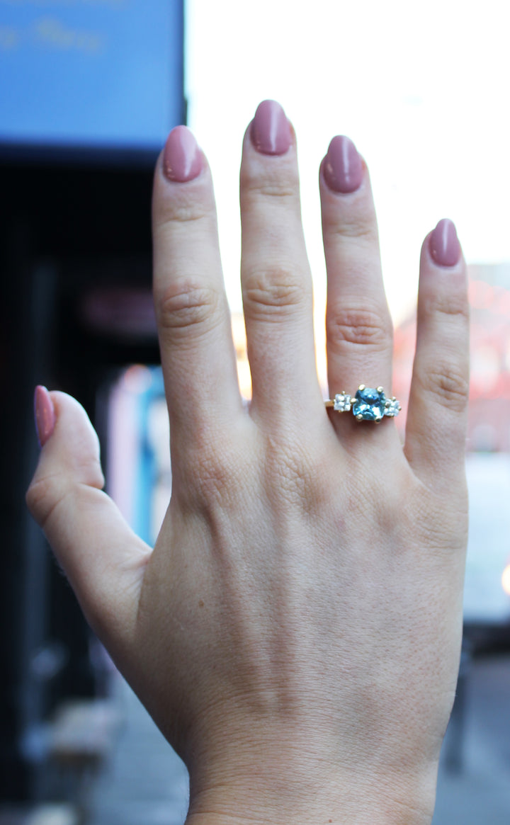 A hand with mauve-painted nails showcases the Madeleine Sky's Devotion 18ct Yellow Gold Ring, featuring an aquamarine center stone flanked by two clear stones. The ring is worn on the ring finger against a blurred urban outdoor backdrop.