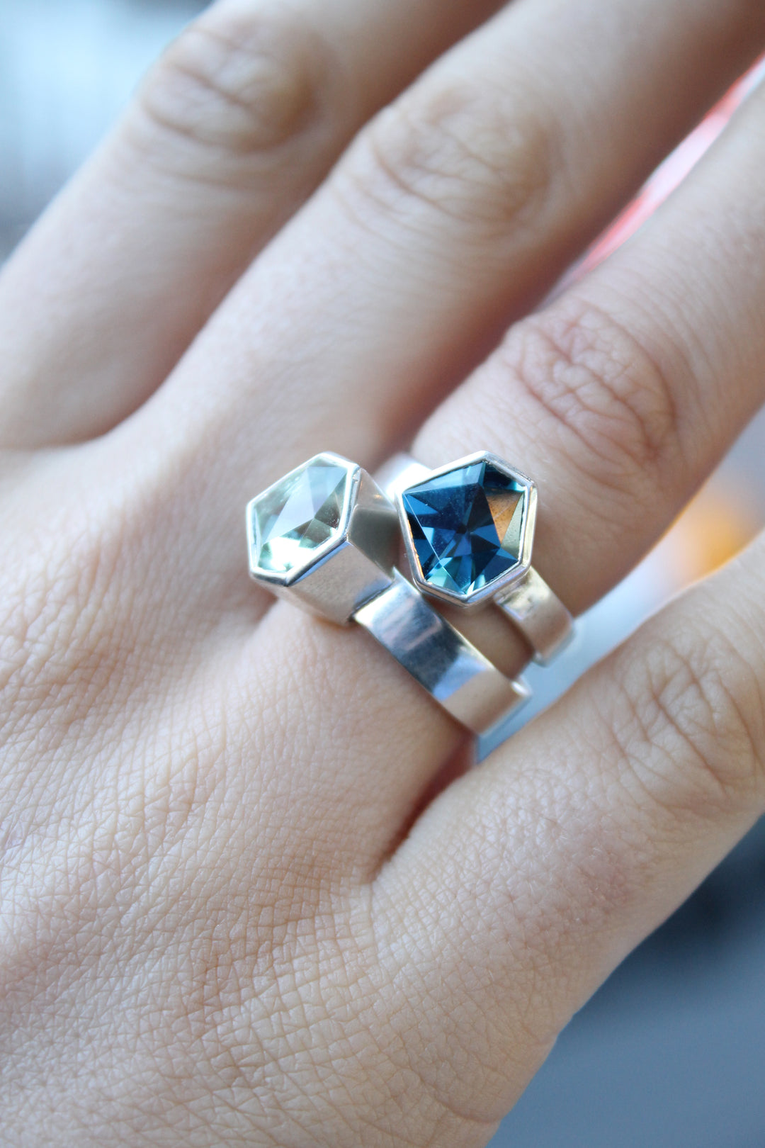 two rings, the first one, closer to the hand is a Silver ring with large, irregular hexagonal green amethyst in a silver bezel. Band is flat and 5mm wide.The second is a Silver ring with large, irregular hexagonal blue topaz in a silver bezel. Band is flat and 4mm wide