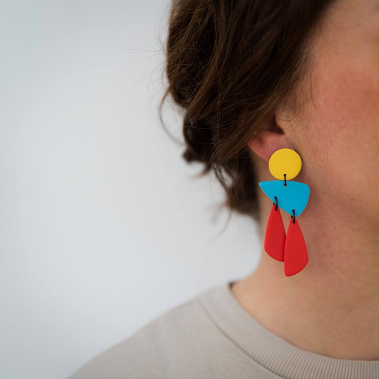 A close-up of a person with brown hair tied back wearing the Taki Primary Earrings by Culu Design—handmade geometric statement earrings in yellow, blue, and red polymer clay. The person is dressed in a light-colored top.
