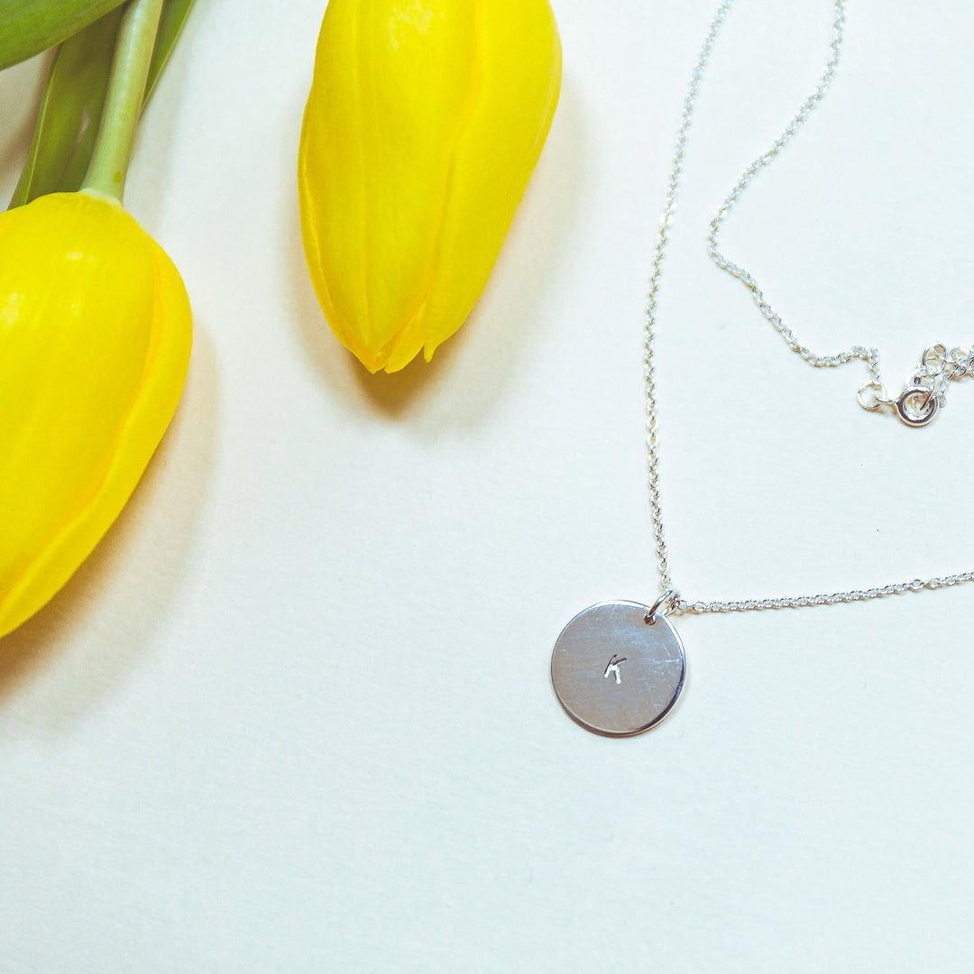 The Hand-stamped Initial Necklace by Personalised Charms, featuring a gold plated sterling silver round pendant, is displayed on a white surface beside two vibrant yellow tulips.
