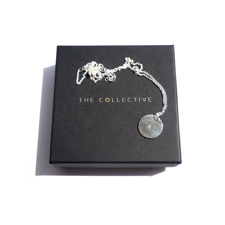 The Hand-stamped Initial Necklace by Personalised Charms, a gold plated sterling silver piece with a round pendant engraved with "1", is displayed on a black box featuring "THE COLLECTIVE" in gold lettering against a white background.