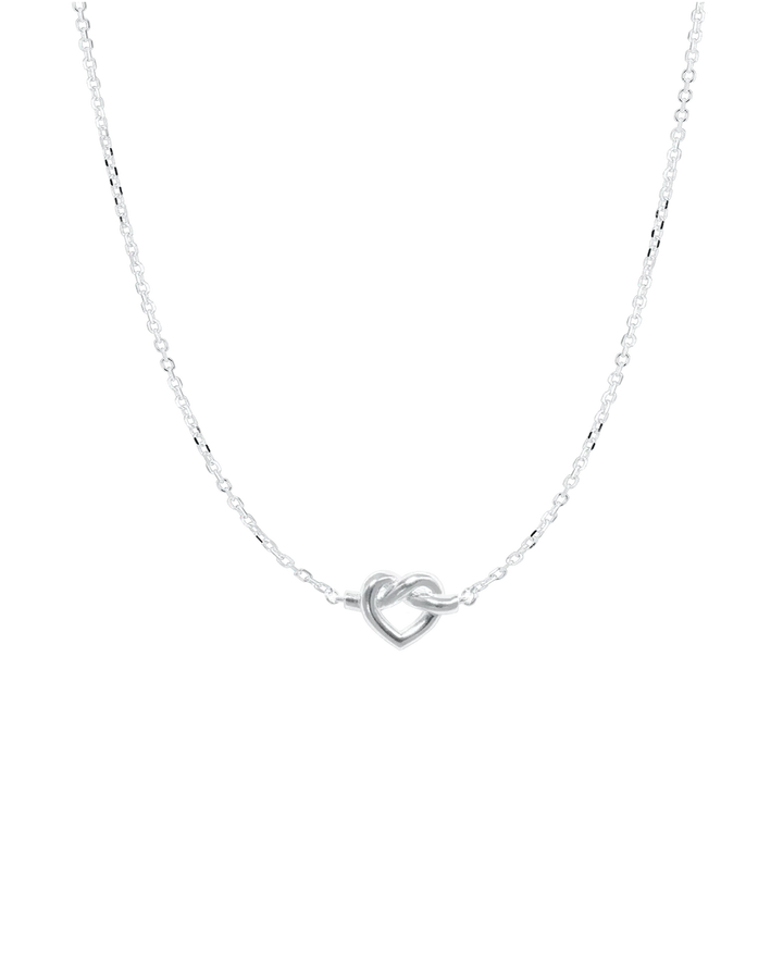 The Tied Heart Necklace by Jane Koenig is a delicate sterling silver piece with a small heart-shaped pendant on an adjustable fine chain, showcasing minimalist elegance against a plain white background.