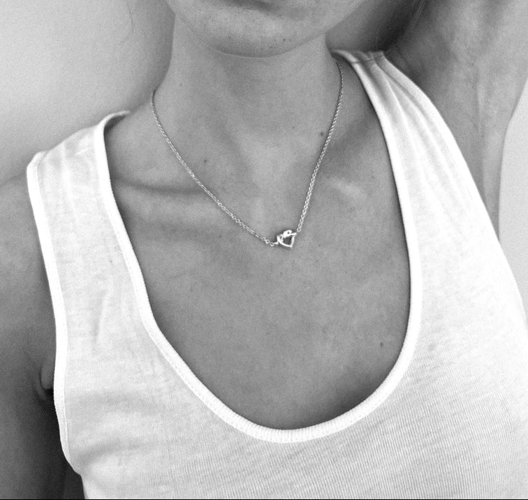 Black and white photo featuring a person in a white tank top, wearing the Jane Koenig Tied Heart Necklace—a delicate sterling silver piece with a small heart pendant. Only the neck, upper chest, and part of the face are shown.
