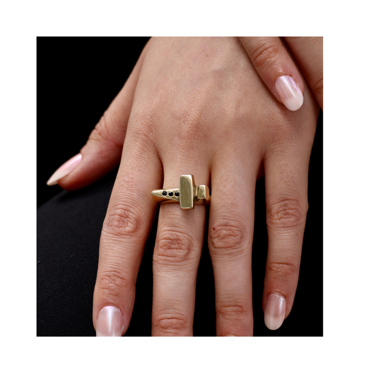 A neatly manicured hand wears the Totem Ring by Madeleine Sculptural Jewellery, a unique hand-carved gold piece shaped like a small hammer with black gemstone accents, elegantly displayed against a black background.