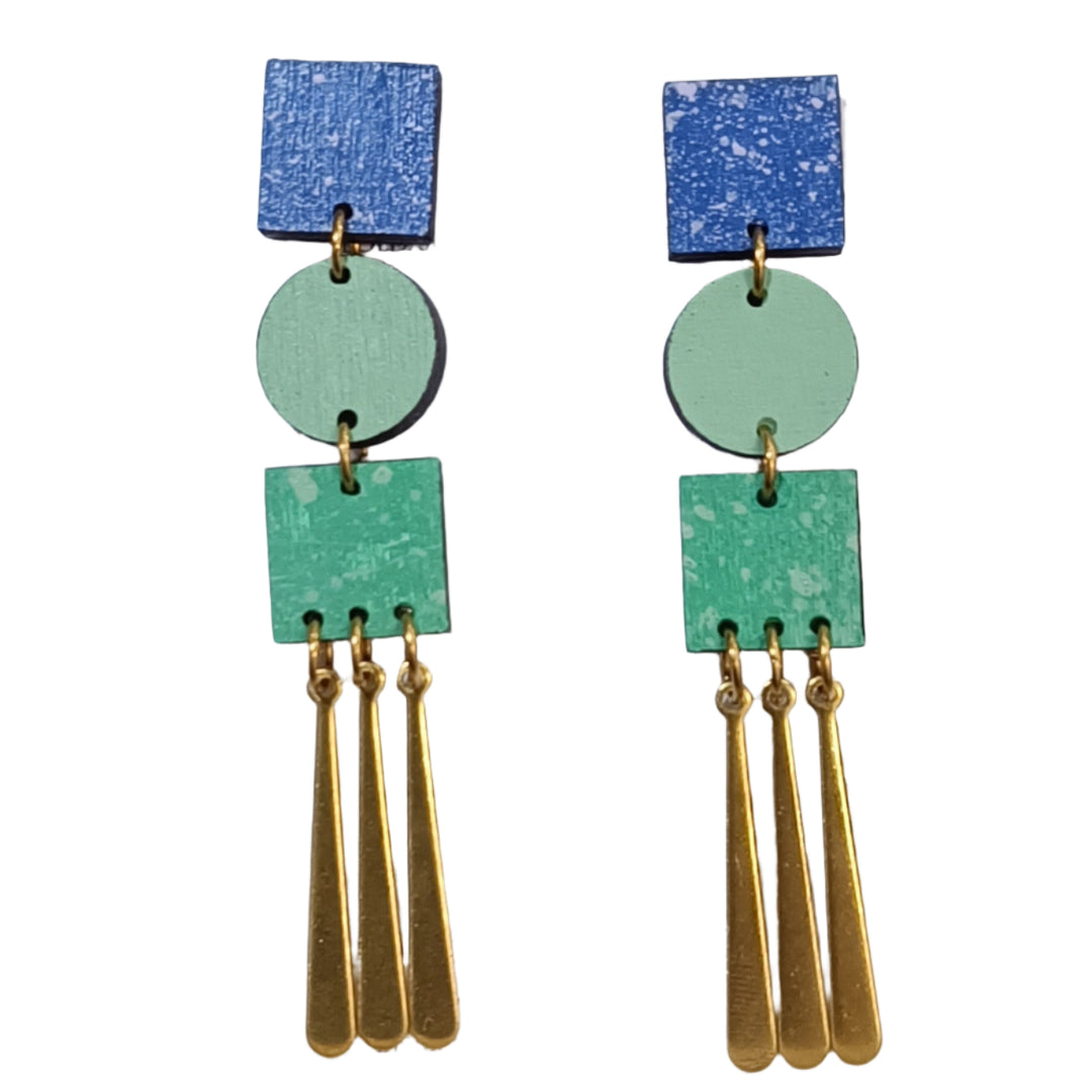The Trio Grande Earrings in Blue & Green by Shock of Grey feature handcrafted birchwood with blue squares, green circles and squares, plus three gold dangling bars, all arranged vertically. Each pair comes in a stylish gift box.