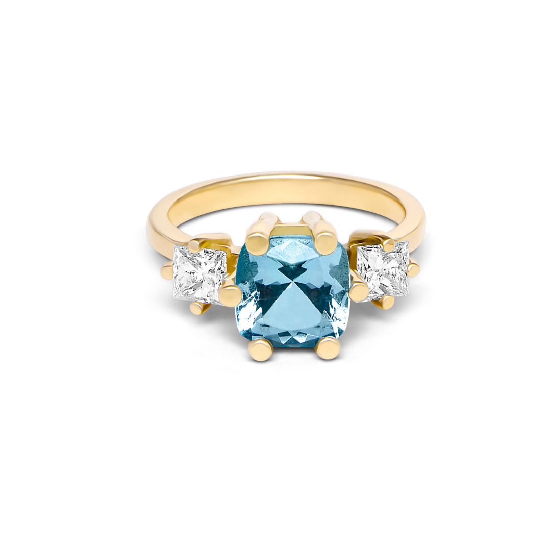 The Sky's Devotion 18ct Yellow Gold Ring by Madeleine showcases a large square-cut blue gemstone and two smaller square-cut clear stones on each side, evoking the elegance of an aquamarine engagement ring.