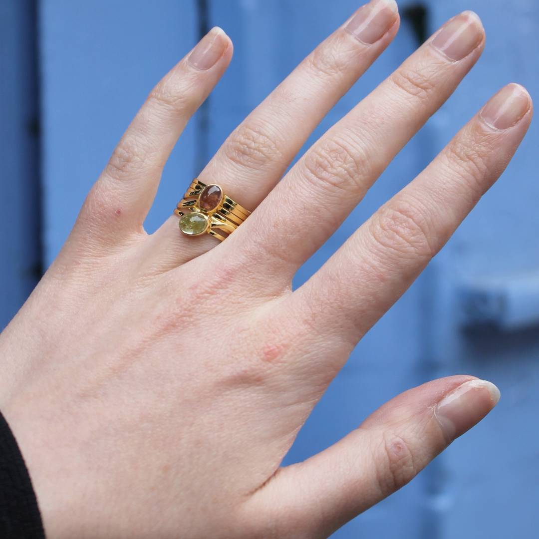 A hand with manicured nails wears three gold rings, including Watermelon tropical's Green and Pink Tourmaline in a Multi Band Gold Ring, held against a light blue background.