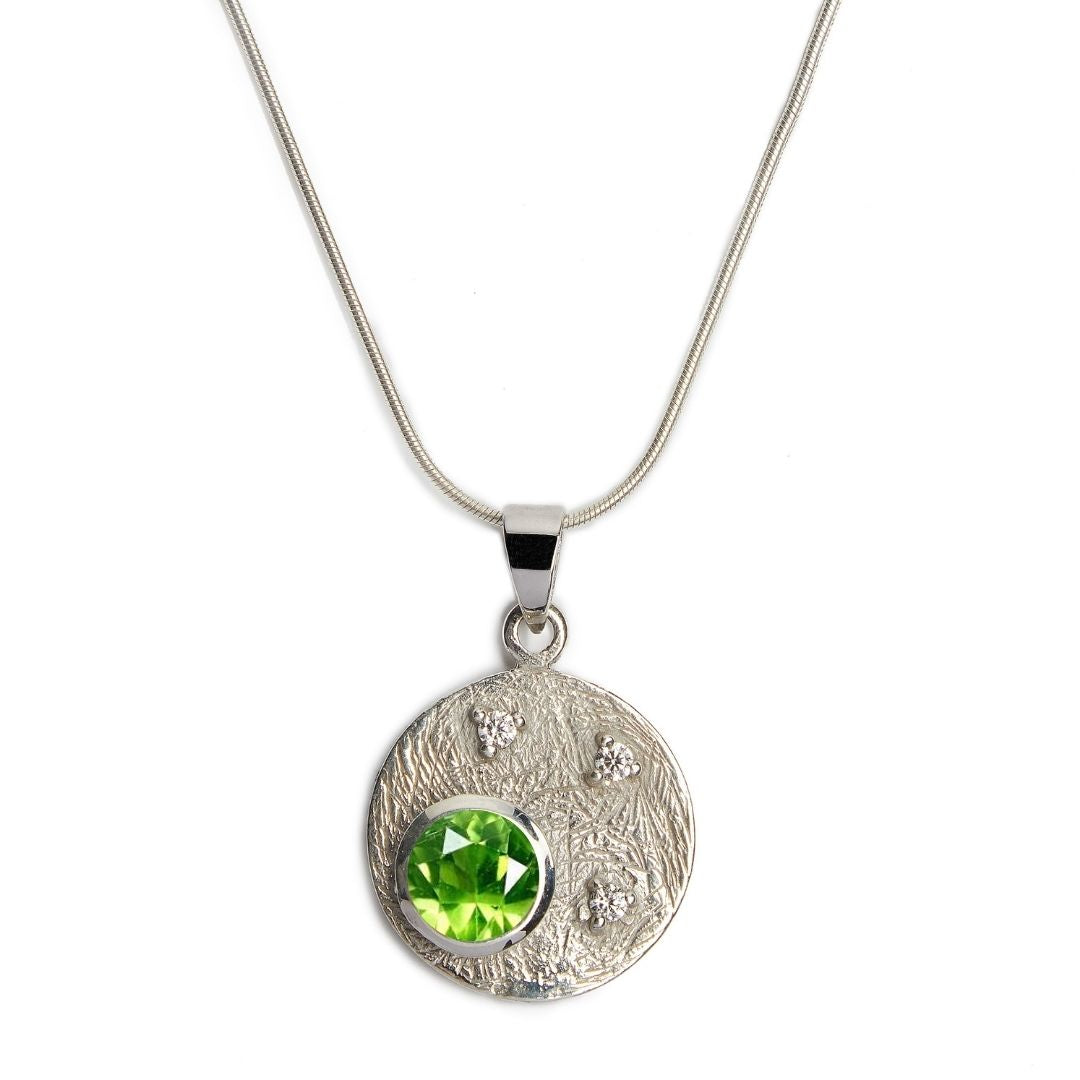 The Celeste Necklace by Gallardo & Blaine Designs features a round textured sterling silver pendant with a large green gemstone and three small cubic zirconia stones.