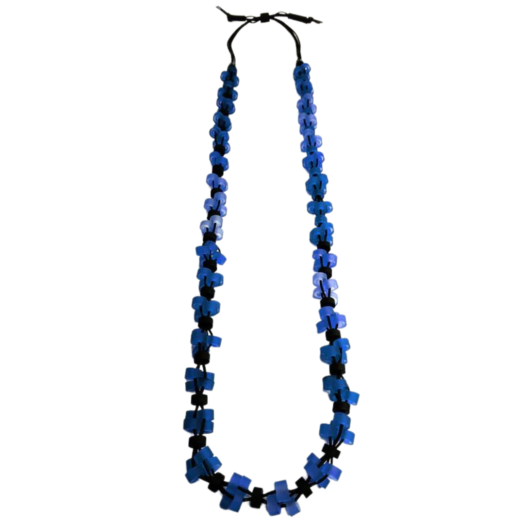 The Alex + Svet Verona Long Necklace is a handmade, hypoallergenic necklace composed of multiple blue cross-shaped beads arranged in a gradient from light blue at the top to dark blue at the bottom, strung on a black cord. The adjustable rubber necklace is long and has a rustic, artisanal appearance.