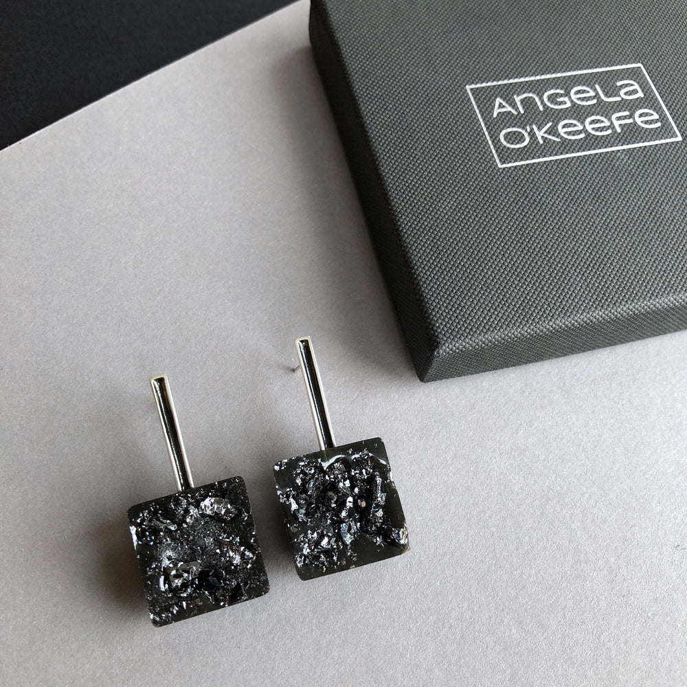 A pair of Angela O'Keefe Urban Geode Square Black Resin Earrings with black textured square stones and sleek metal stems is shown on a gray and black surface. To the right, there is a dark gray box with the name "Angela O'Keefe" written on the lid in white text.